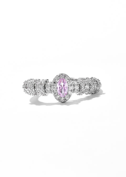 An oval ring with a central pink gemstone surrounded by tiny clear stones set in a gold plated band.