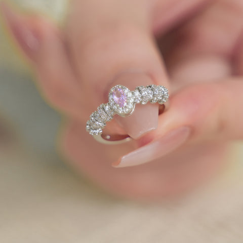 An oval ring with a central pink gemstone surrounded by tiny clear stones set in a gold plated band.