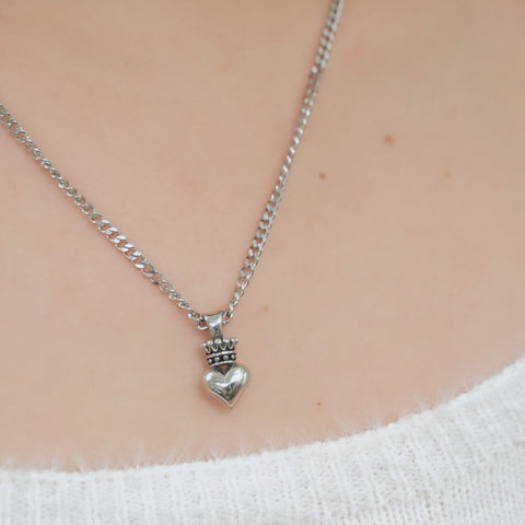 A silver chain necklace, holding a heart pendant topped with a small crown.